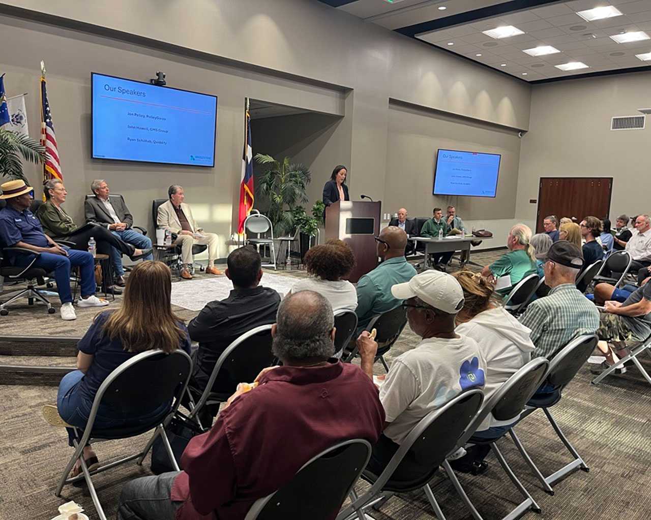 Speaker at a podium addresses an audience during the Bridgestone MUD State of the District meeting. Panelists sit on stage next to a presentation screen and flags.