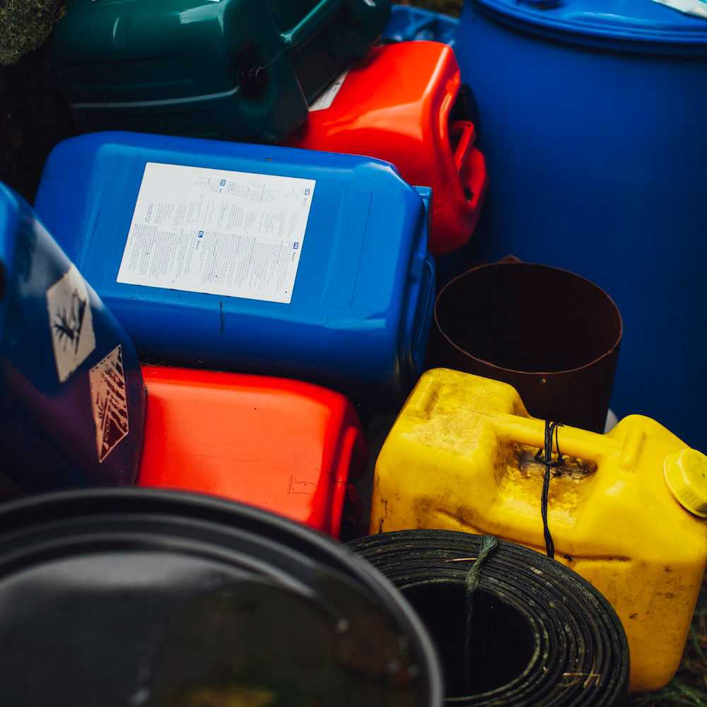 Various colorful industrial containers, including blue, red, and yellow plastic jugs and barrels, stacked together with hazard warning labels visible on their sides.