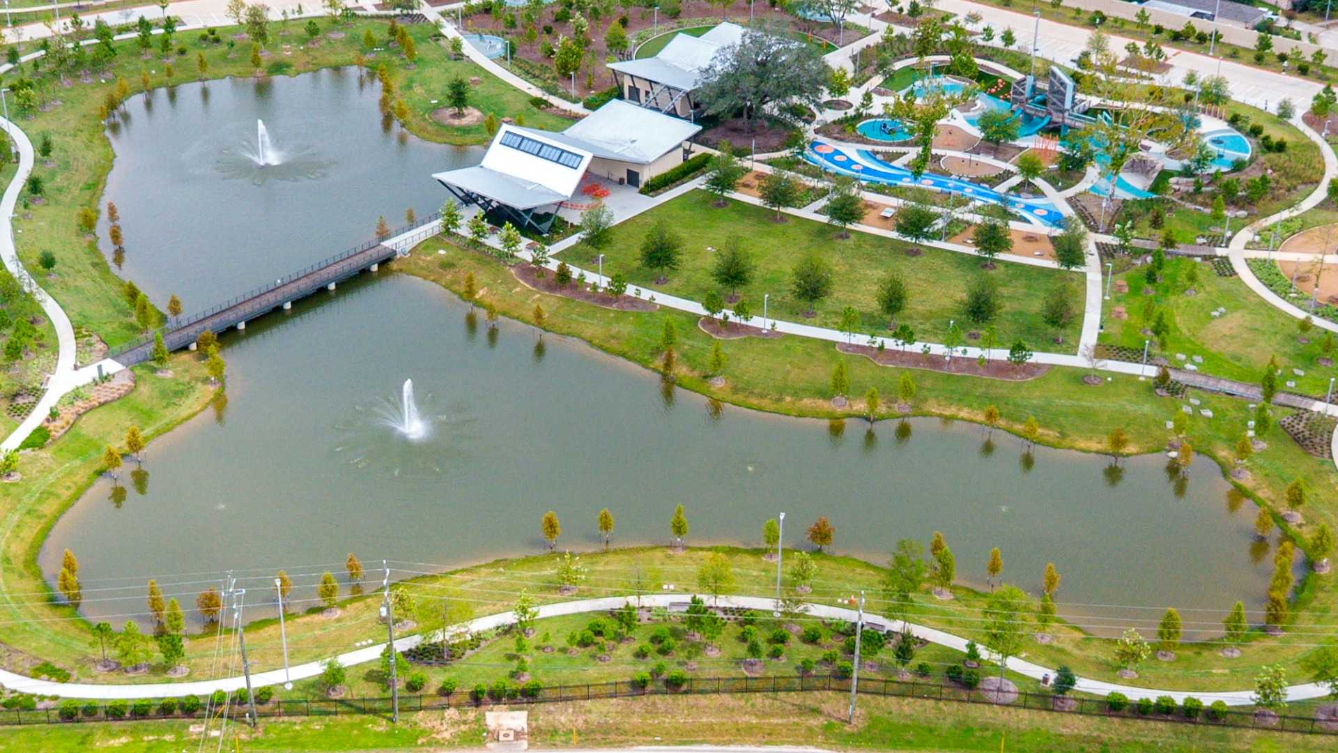 An aerial view of a modern community park featuring a large pond with two fountains, a wooden pedestrian bridge, paved walking trails, a colorful playground, and contemporary pavilion buildings.