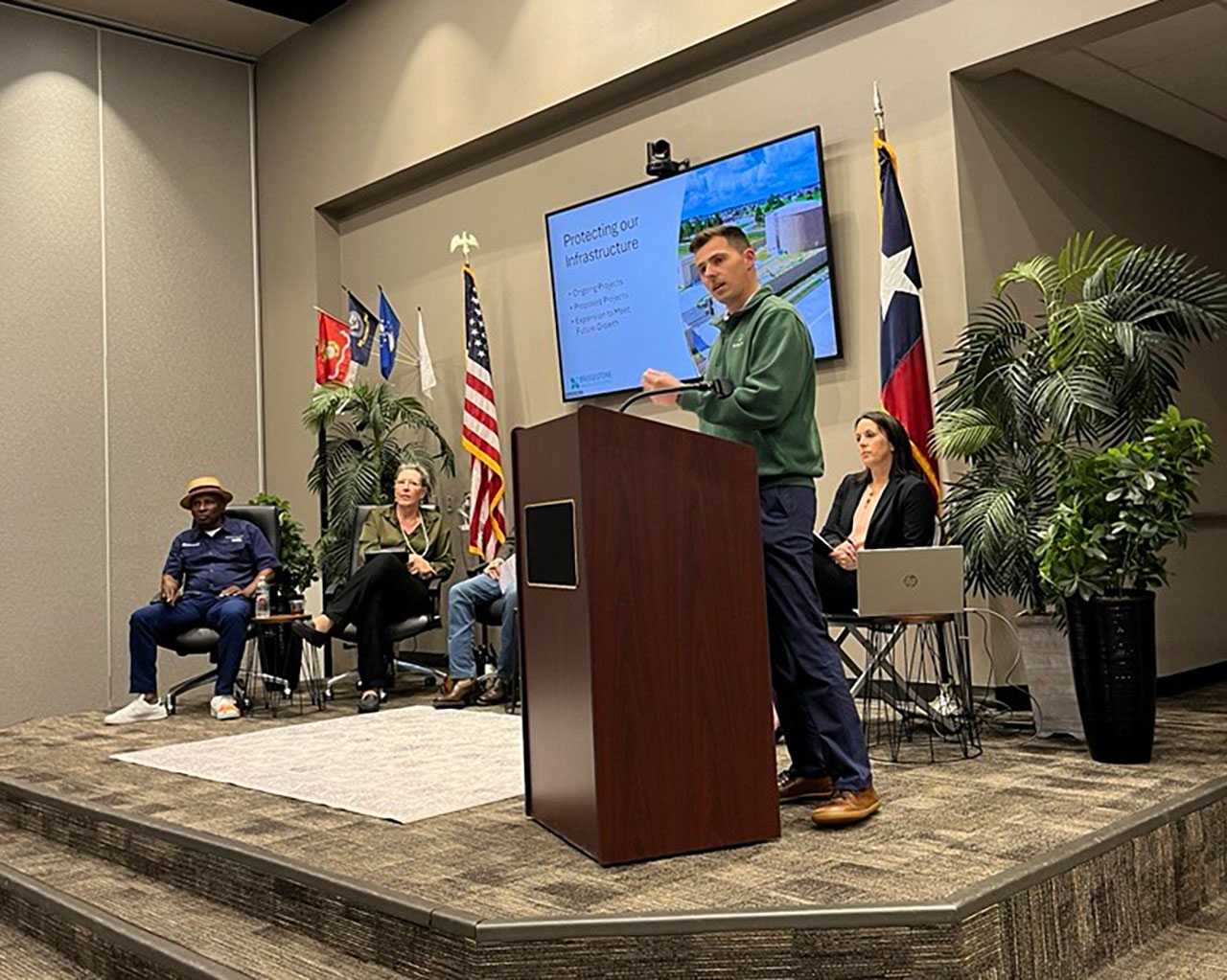 Speaker at a podium presents a Protecting our Infrastructure slideshow during the Bridgestone MUD State of the District meeting. Panelists sit on stage flanked by American and Texas flags.