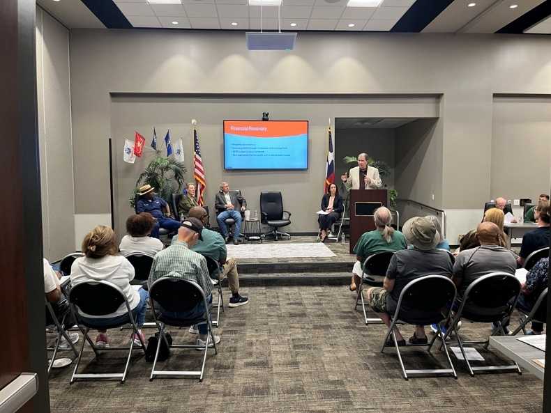 A speaker at a podium presents a financial review slideshow to an audience in a community meeting room. Panelists sit on stage behind him, flanked by American and Texas flags.